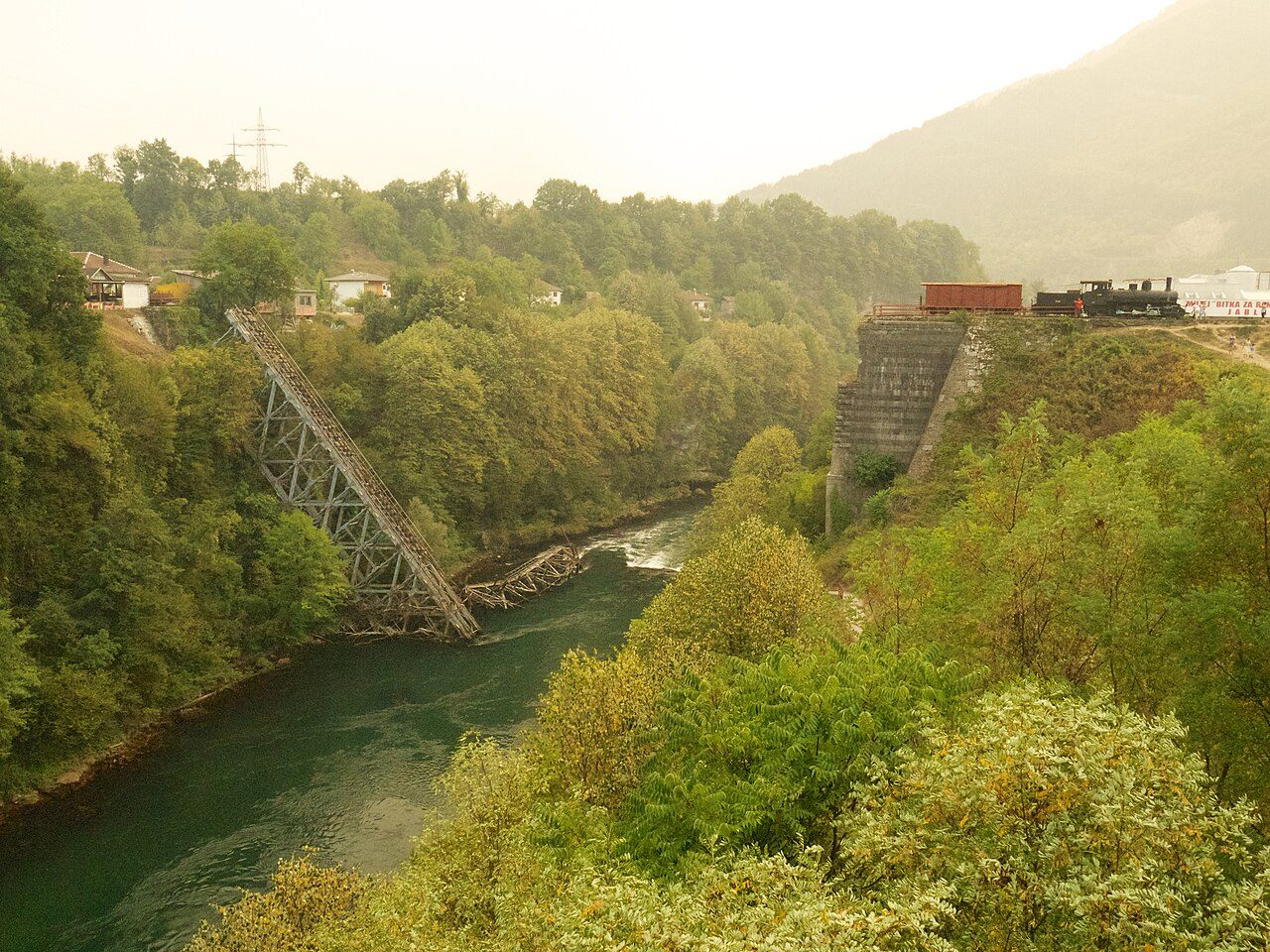 The collapsed railway bridge hanging into the Neretva at Jablanica, with an old train on the bank above