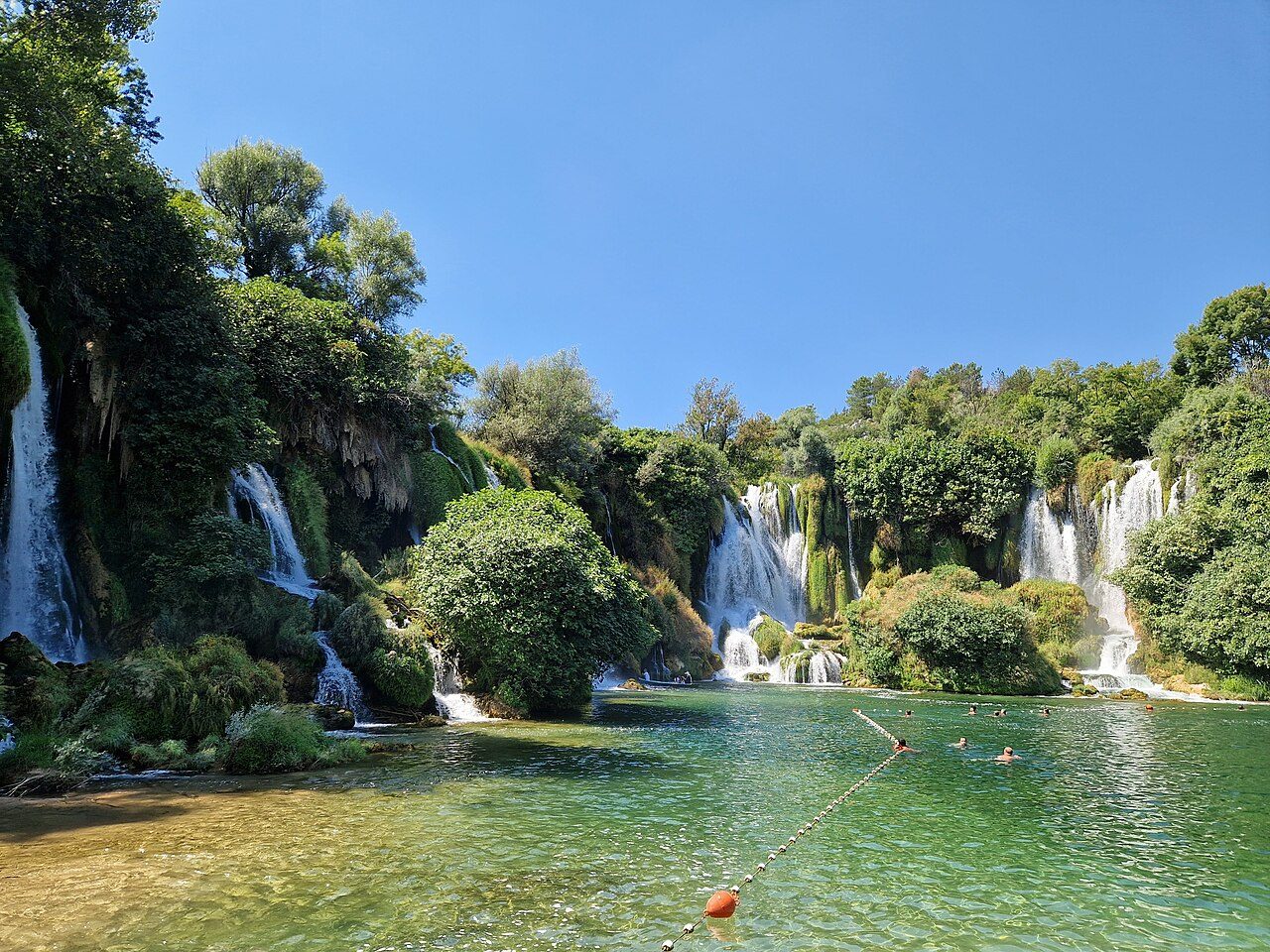 The Kravica waterfalls pouring into a green pool, surrounded by trees
