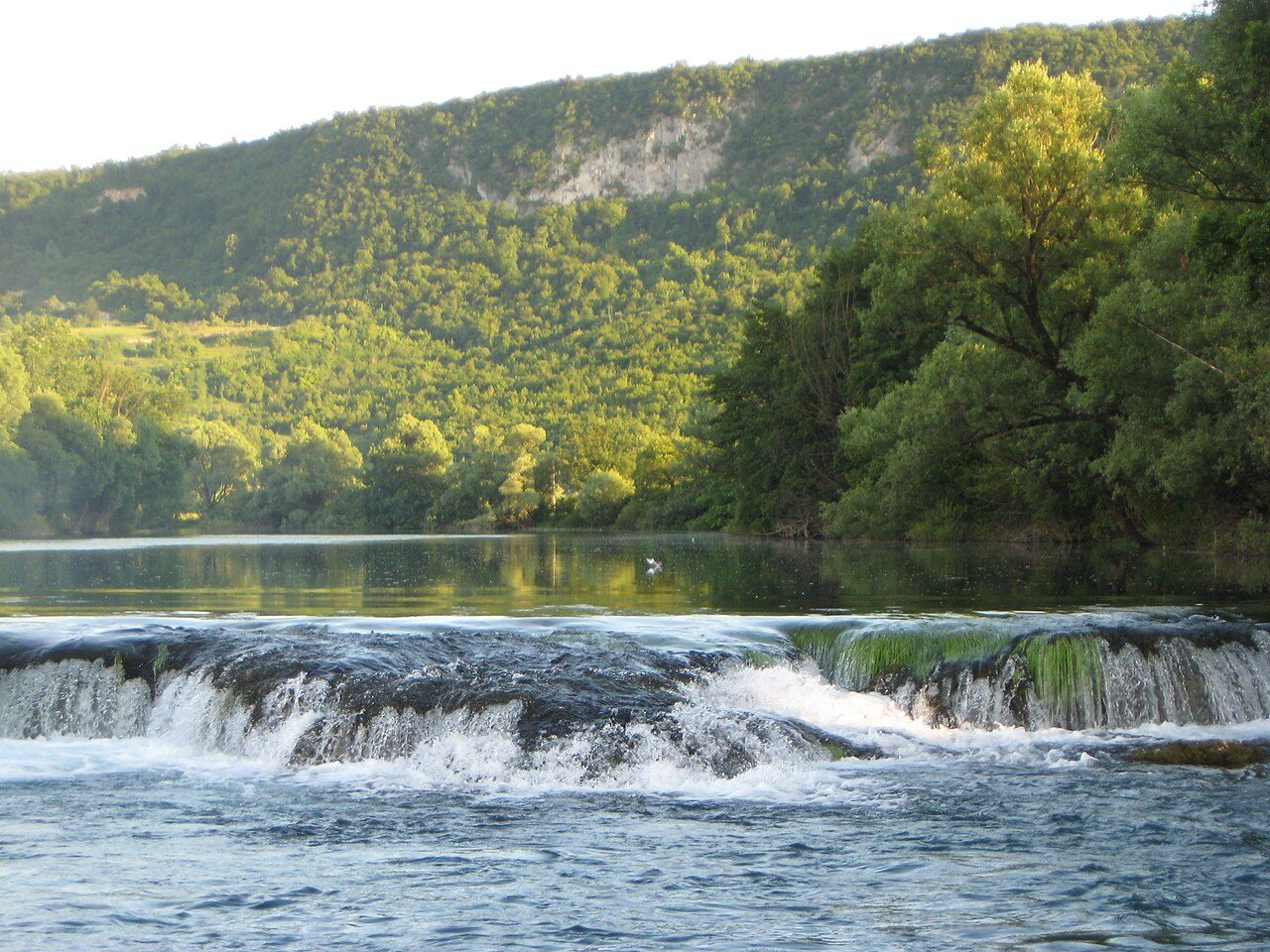 The Una river spilling over a low rocky weir near Golubić, with wooded hills behind